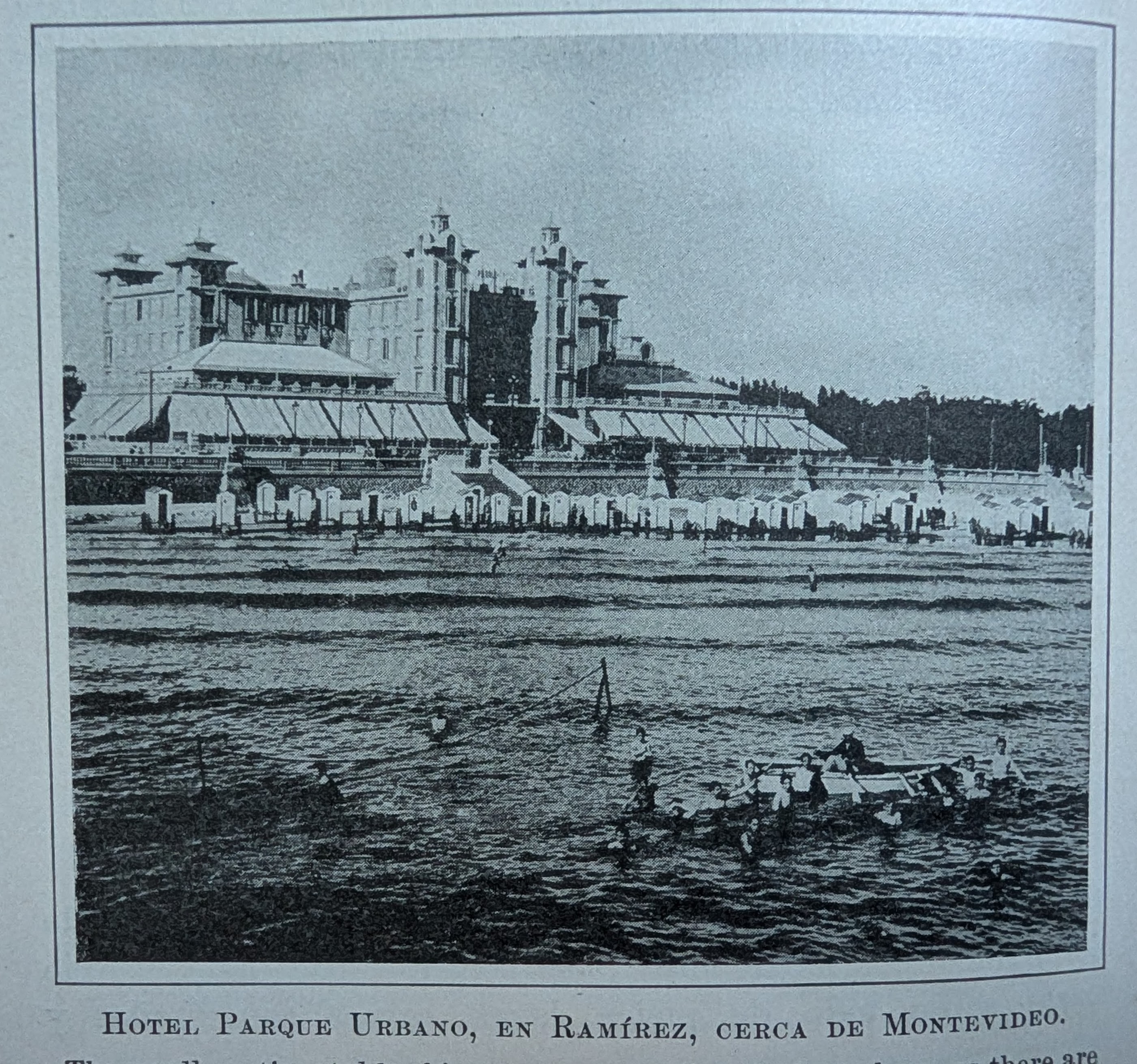 Antique photo taken from the ocean of people wading around a boat and changing tents lining the shoreline. In the background is a hotel. The caption, in Spanish, reads, Urbano Park Hotel, in Ramirez, near Montevideo.