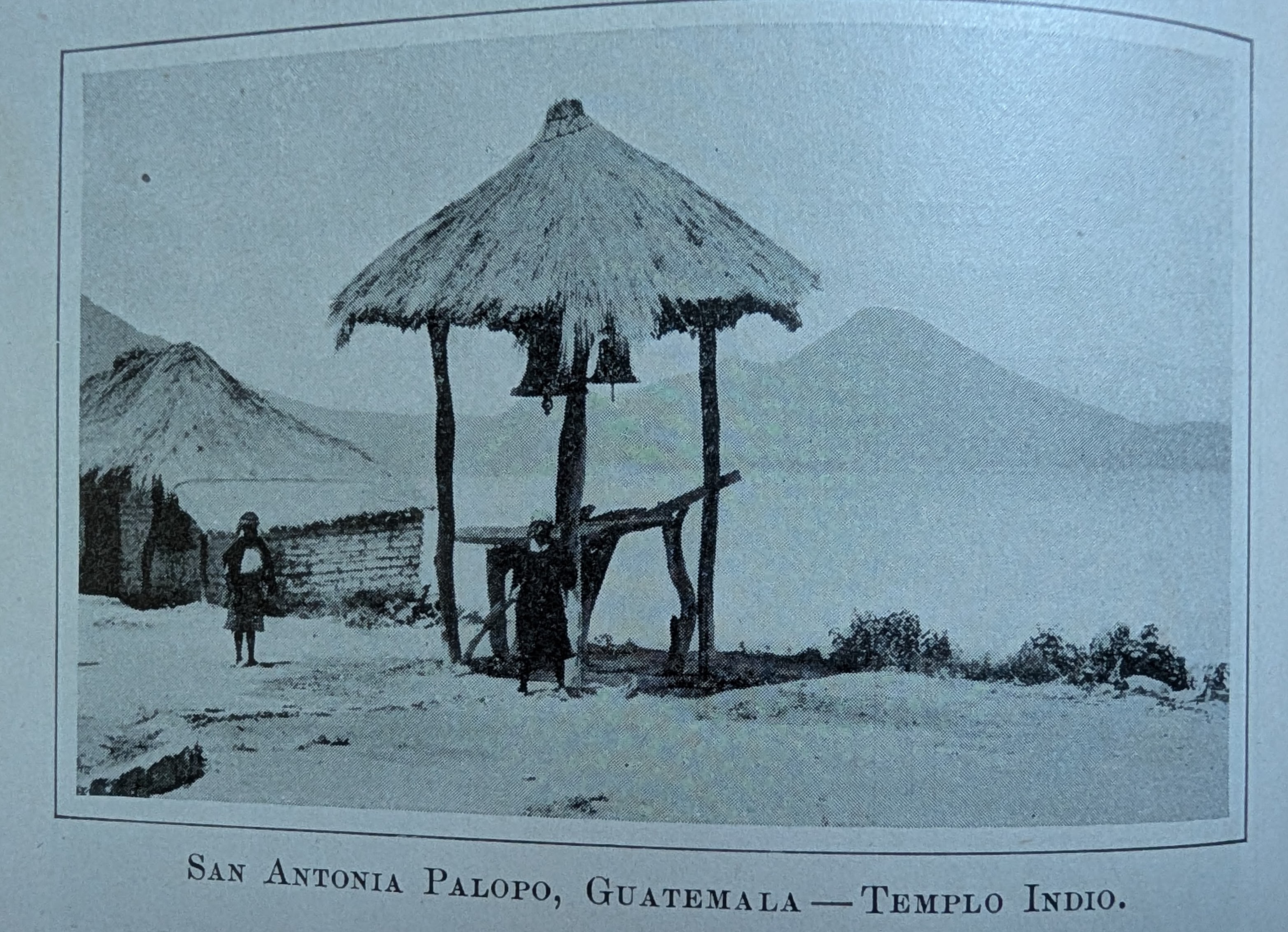 Antique photo showing two people in front of a wooden and straw belltower and clay brick building. Behind them are mountains and a lake. The caption, in Spanish, reads, San Antonia Palopo, Gatamala, Indian Temple.